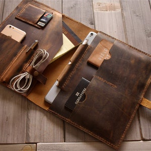 May include: A brown leather portfolio with multiple pockets and compartments. The portfolio is open and shows a tablet, passport, phone, and other items.