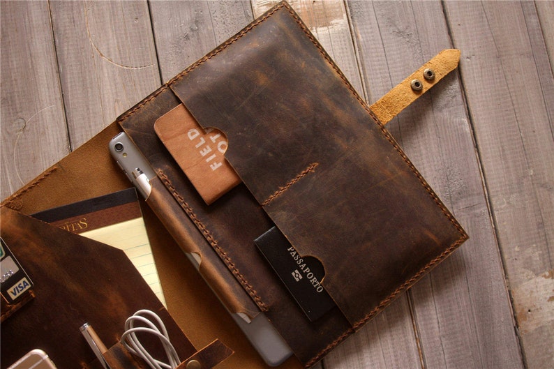Op de afbeelding: Een bruine leren portfolio met meerdere zakken en vakken. De portfolio is open en bevat een tablet, een notitieboek, een pen, een paspoort en een creditcard. De portfolio heeft een bruine leren band met twee metalen drukknopen.