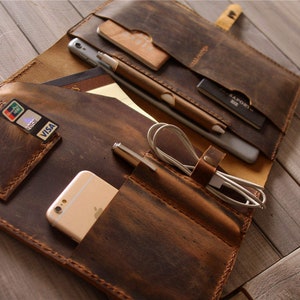 Op de afbeelding: Een bruin leren portfolio met meerdere vakken en slots om items zoals een telefoon, pennen, kaarten en een tablet te dragen. De portfolio is open en het interieur is zichtbaar.