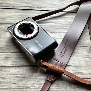 Può includere: Un cinturino per fotocamera in pelle marrone con il testo "The Best scenery is at road, just start off today. ZZ" inciso su di esso. Il cinturino è attaccato a una fotocamera Samsung nera.