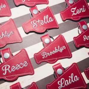 May include: Red leather megaphone-shaped keychains with white embroidered names, including Stella, Brooklyn, Reece, Laila, and Zeni.