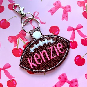 May include: A brown, textured football-shaped keychain featuring white embroidered laces and the name "kenzie" stitched in bright pink thread. The keychain includes a silver-toned metal lobster clasp and a circular ring attached with a white snap button.