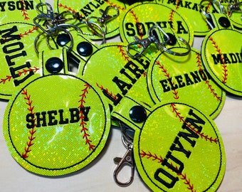 May include: A close-up of several green and white softball-shaped keychains with different names embroidered on them. The keychains have a black button and a silver metal clasp.