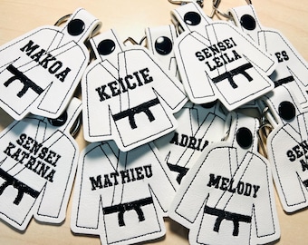 May include: A collection of white karate uniform keychains with black embroidered belts and names like Makoa, Keicie, Sensei Leila, Sensei Katrina, Mathieu, Adri, Melody, and Sensei Ald.