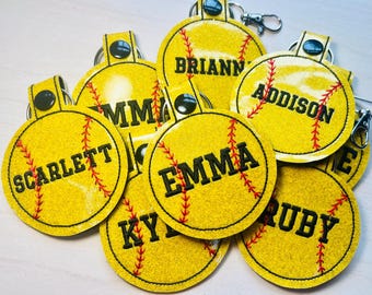 May include: A collection of round, glittery yellow softball keychains featuring red stitched seams and black embroidered names including Scarlett, Emma, Briann, Addison, and Ruby. Each keychain has a black snap button and a metal lobster clasp.