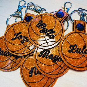 May include: A set of seven gold glitter basketball keychains with black stitching and personalized names. The names are "Jozy", "Wyllo", "Lula", "May", "Pas", "Nu", and "Jozy".