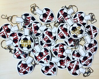 May include: A pile of white heart-shaped keychains with black and white soccer ball designs. Each keychain has a name and number embroidered on it, including "Coach Matt" and "Coach Joe".