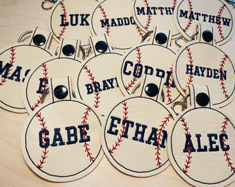 May include: A collection of white leather baseball-themed keychains with red stitching and blue embroidered names. The names include:  Luke, Maddox, Matthew, Hayden, Ethan, Alec, Gabe, Braylon, Mason, and Corden.