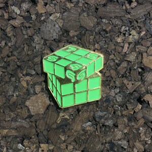 May include: A green and gold metal pin shaped like a Rubik's cube with the letter "S" on each side.