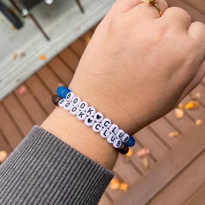 May include: Two beaded bracelets with the words "BOOK CLUB" spelled out in white letter beads. The bracelets have blue and brown end beads. A gold ring with a large, clear stone is also visible.