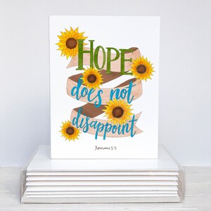 May include: A white greeting card featuring the words "HOPE does not disappoint" in green and blue script, with a brown ribbon and yellow sunflowers. The text "Romans 5:5" is at the bottom.