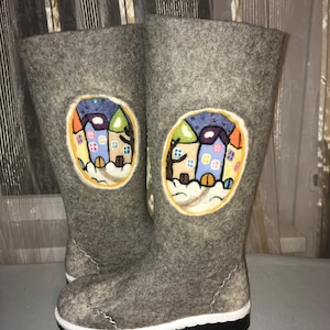 Puede incluir: Un par de botas de fieltro gris con suela blanca. Las botas tienen un diseño bordado de colores de una casa con techo amarillo y ventanas azules.
