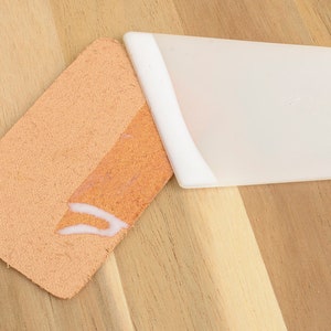 May include: A close-up of a light orange leather piece with a white adhesive applied to it, next to a white plastic spatula. The items are on a light brown wooden surface.
