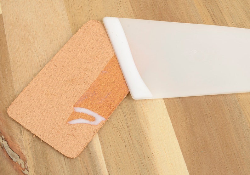 May include: A close-up of a light orange leather piece with a white adhesive applied to it, next to a white plastic spatula. The items are on a light brown wooden surface.