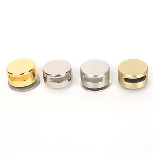 May include: Four metallic bottle stoppers in gold, silver, and brass are displayed on a white background. Each stopper has a circular design with a flat top and a small, horizontal slot. These stoppers are likely for wine or other bottles.
