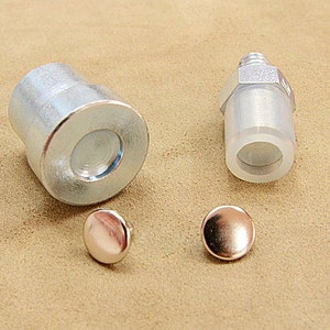 May include: An assortment of metal hardware components, including a cylindrical piece, a threaded fitting, and two small, round, copper-colored rivets. The items are displayed on a neutral, tan-colored surface.