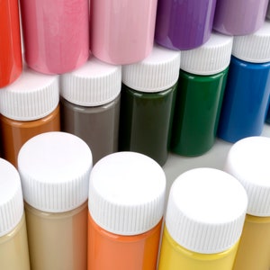 May include: A collection of small, cylindrical paint bottles in various colors, including red, pink, purple, green, blue, and yellow. Each bottle has a white cap. The bottles are arranged closely together, showcasing a spectrum of hues.