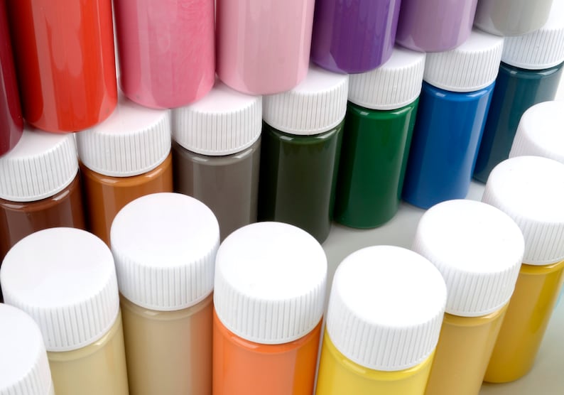 May include: A collection of small, cylindrical paint bottles in various colors, including red, pink, purple, green, blue, and yellow. Each bottle has a white cap. The bottles are arranged closely together, showcasing a spectrum of hues.