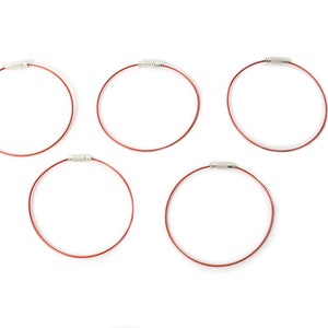 May include: Five red wire key rings with silver-colored clasps are arranged against a white background. The key rings are circular and appear to be made of a flexible metal wire. The clasps are small and cylindrical.