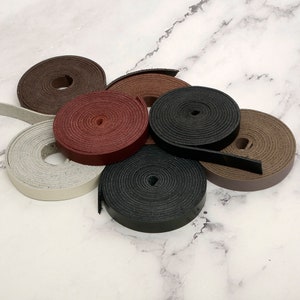 May include: A collection of leather strips in various colors, including brown, black, and gray, are coiled into neat rolls. The leather appears to be smooth and of high quality, suitable for crafting or other projects. The rolls are arranged on a light-colored surface.