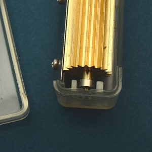 Puede incluir: Primer plano de un mecanismo de caja de m&uacute;sica. La imagen muestra un peine dorado y un engranaje en forma de estrella. El mecanismo est&aacute; alojado en una caja de pl&aacute;stico transparente, con un tornillo visible en el lateral. El fondo es de color verde azulado oscuro.