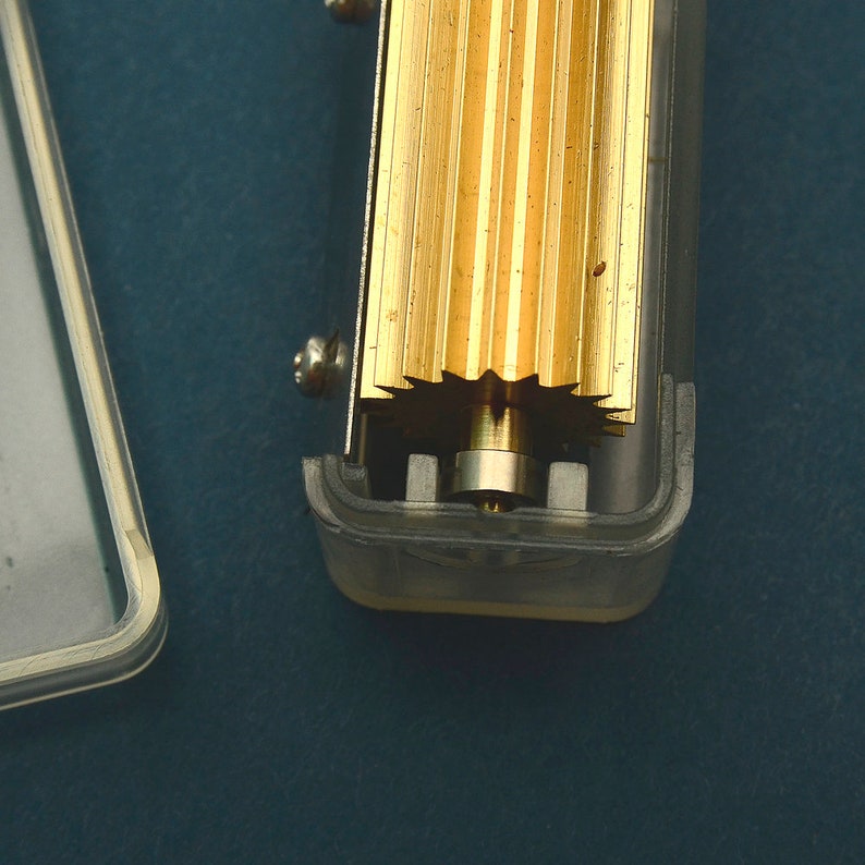 Puede incluir: Primer plano de un mecanismo de caja de m&uacute;sica. La imagen muestra un peine dorado y un engranaje en forma de estrella. El mecanismo est&aacute; alojado en una caja de pl&aacute;stico transparente, con un tornillo visible en el lateral. El fondo es de color verde azulado oscuro.