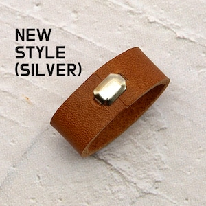 Peut inclure: Un bracelet en cuir marron avec un accent rectangulaire argenté. Le texte "NEW STYLE (SILVER)" est en haut à gauche. Le bracelet est de conception simple et moderne.