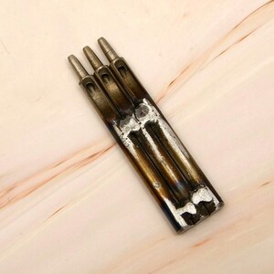 Puede incluir: Una herramienta de punzonado de cuero metálico con tres punzones cilíndricos. La herramienta tiene un cuerpo rectangular con una construcción soldada y un acabado metálico desgastado. Las puntas de los punzones son ligeramente cónicas.