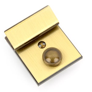 Könnte beinhalten: Rechteckiger, messingfarbener Verschluss mit gebürsteter Oberfläche. Der Verschluss hat einen kleinen, runden, dunkel getönten Knopf und einen rechteckigen Bügel. Die Hardware ist wahrscheinlich für eine Tasche oder ein anderes Accessoire.