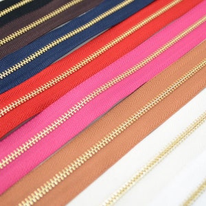 Puede incluir: Una colecci&oacute;n de cremalleras coloridas con dientes de metal dorado. Las cremalleras son de varios colores, incluyendo negro, marr&oacute;n, azul marino, rojo, rosa, marr&oacute;n claro y blanco. La imagen es un primer plano.