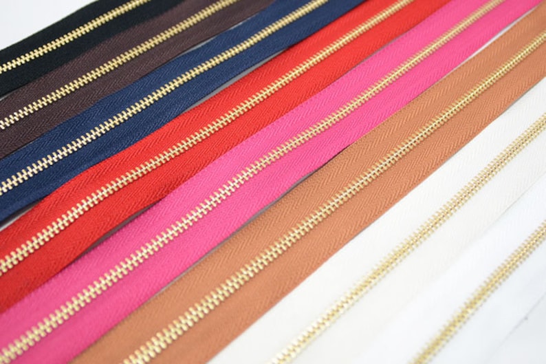 Puede incluir: Una colecci&oacute;n de cremalleras coloridas con dientes de metal dorado. Las cremalleras son de varios colores, incluyendo negro, marr&oacute;n, azul marino, rojo, rosa, marr&oacute;n claro y blanco. La imagen es un primer plano.