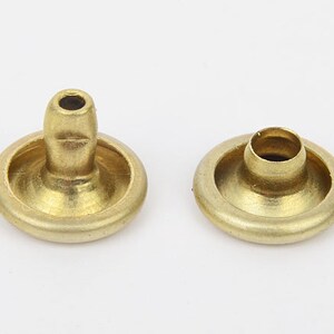 Puede incluir: Dos remaches de latón, con cabeza redondeada y vástago cilíndrico. Los remaches son de color dorado cálido y están diseñados para sujetar materiales. Comúnmente utilizados en marroquinería y confección.