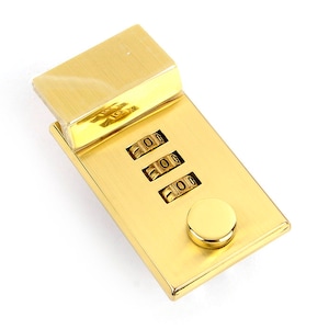 May include: A gold-coloured combination lock with three number dials and a small circular button. The lock has a rectangular shape with a hinged top. The numbers on the dials are visible.