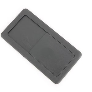 May include: A dark grey rectangular plastic item with a recessed section and a sliding cover. The cover is slightly raised and has a small, curved indentation. The item is set against a white background.