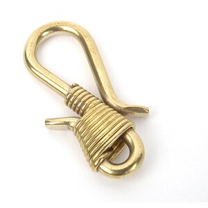 May include: A brass key hook with a curved top and a spring-loaded clasp. The hook is wrapped with coiled brass wire for added detail. The key hook is designed to attach to a belt loop or bag.