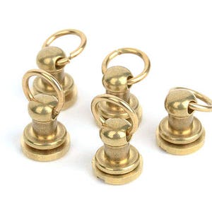 May include: A collection of five brass-colored rivets with attached circular rings. Each rivet has a rounded top and a flat, circular base. The rivets are arranged against a white background, showcasing their metallic sheen.