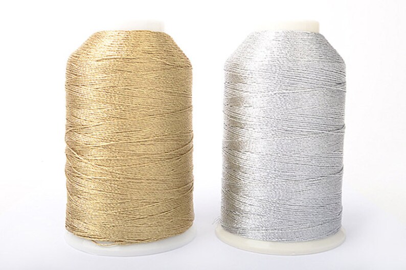 May include: Two spools of metallic thread. One spool is gold, the other is silver. The thread is tightly wound around a white plastic base. The spools are side by side against a white background. Ideal for embroidery and sewing projects.