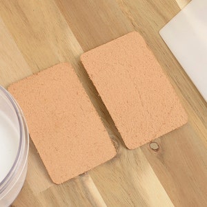 May include: Two rectangular, peach-colored leather pieces with a textured surface sit on a wooden surface. A clear container with a white liquid and a white spatula are also present.