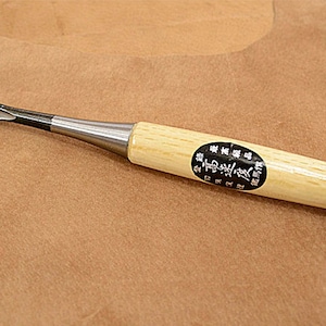 Puede incluir: Un formón de madera con mango de madera clara y hoja de metal. El formón tiene una virola plateada y un extremo metálico redondeado. Una etiqueta en blanco y negro está adherida al mango.