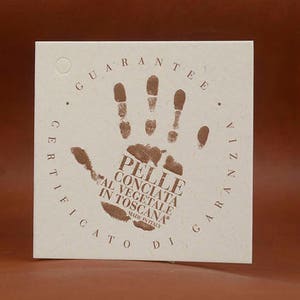 May include: A square, off-white card with a brown handprint and text. The handprint is surrounded by a circular text that reads "GUARANTEE" and "CERTIFICATO DI GARANZIA". The text in the handprint reads "PELLE CONCIATA AL VEGETALE IN TOSCANA MADE IN ITALY".