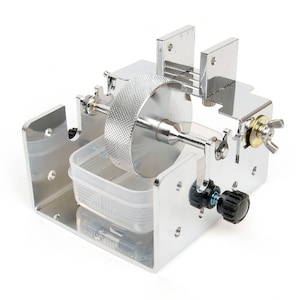 Pode incluir: Uma máquina de metal prateado com uma grande roda texturizada no centro. Possui um recipiente de plástico transparente e botões ajustáveis. A máquina é projetada para um propósito específico, provavelmente para artesanato ou fabricação em pequena escala.