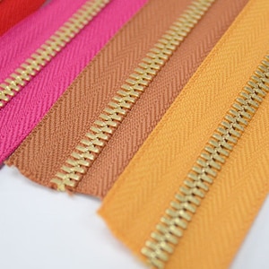 Puede incluir: Primer plano de cremalleras coloridas en rojo, rosa, marr&oacute;n y naranja. Cada cremallera tiene un tirador de metal dorado. La tela presenta un sutil patr&oacute;n de espiga. Adecuado para ropa o proyectos de manualidades.