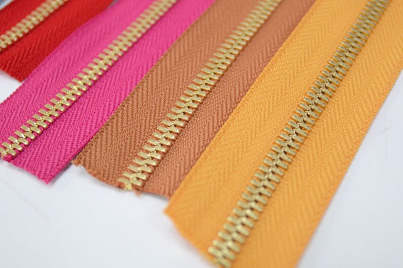 Puede incluir: Primer plano de cremalleras coloridas en rojo, rosa, marr&oacute;n y naranja. Cada cremallera tiene un tirador de metal dorado. La tela presenta un sutil patr&oacute;n de espiga. Adecuado para ropa o proyectos de manualidades.