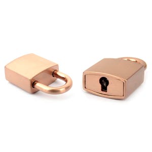 May include: Two rose gold padlocks. One padlock is open, with the U-shaped shackle extended. The other padlock is closed, with a keyhole visible. Both padlocks have a rectangular body and rounded edges.
