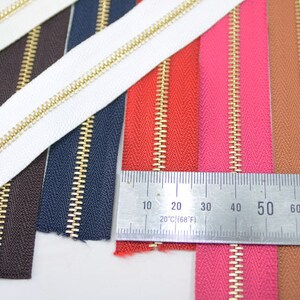 Puede incluir: Primer plano de varias cremalleras de colores con dientes dorados. Las cremalleras son de varios colores, incluyendo blanco, negro, azul marino, marr&oacute;n, rojo, rosa y beige. Una regla se coloca sobre las cremalleras, mostrando medidas en cent&iacute;metros y grados Fahrenheit.