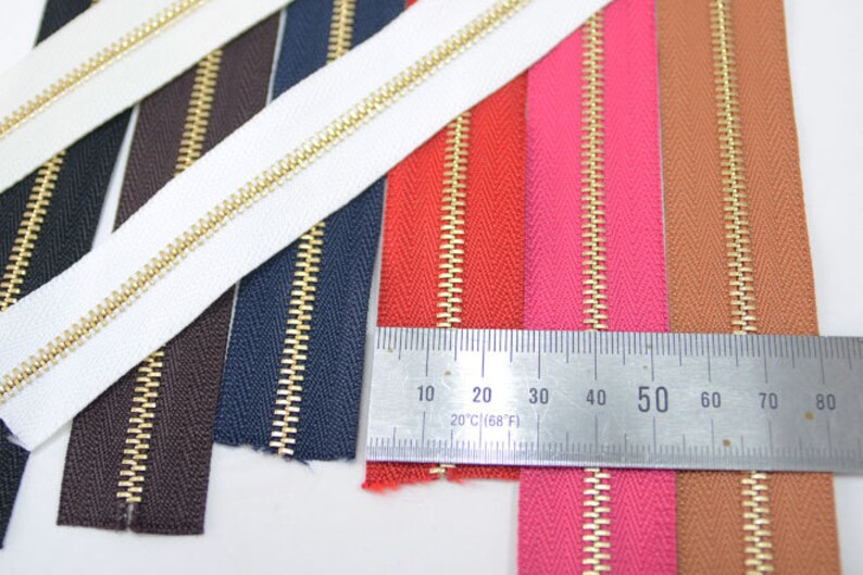 Puede incluir: Primer plano de varias cremalleras de colores con dientes dorados. Las cremalleras son de varios colores, incluyendo blanco, negro, azul marino, marr&oacute;n, rojo, rosa y beige. Una regla se coloca sobre las cremalleras, mostrando medidas en cent&iacute;metros y grados Fahrenheit.