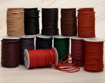 Puede incluir: Una colección de carretes de cordón de cuero trenzado en varios colores, incluyendo beige, negro, marrón, rojo, morado y verde. Los cordones están enrollados firmemente en carretes blancos, mostrando la textura y las variaciones de color. Ideal para manualidades y joyería.
