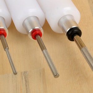 May include: Three white-bodied tools with metal shafts and textured handles. The tools have red and black accents. The shafts have different tip textures. The tools are arranged on a light brown wooden surface.