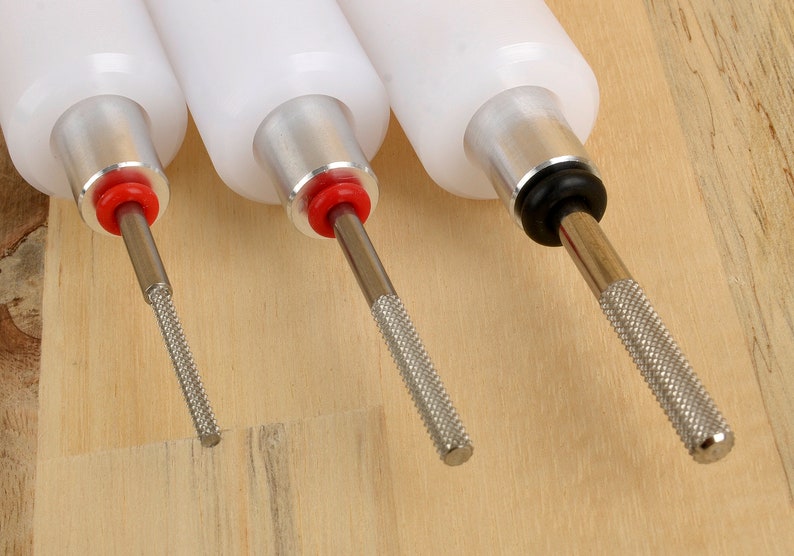 May include: Three white-bodied tools with metal shafts and textured handles. The tools have red and black accents. The shafts have different tip textures. The tools are arranged on a light brown wooden surface.