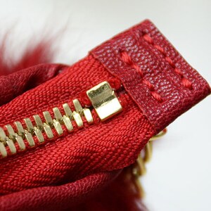 May include: Close-up of a red fabric with a gold zipper. The zipper is partially open, revealing the gold teeth and a gold zipper pull. The fabric has a textured appearance and is stitched with red thread. A small amount of red fur is visible.